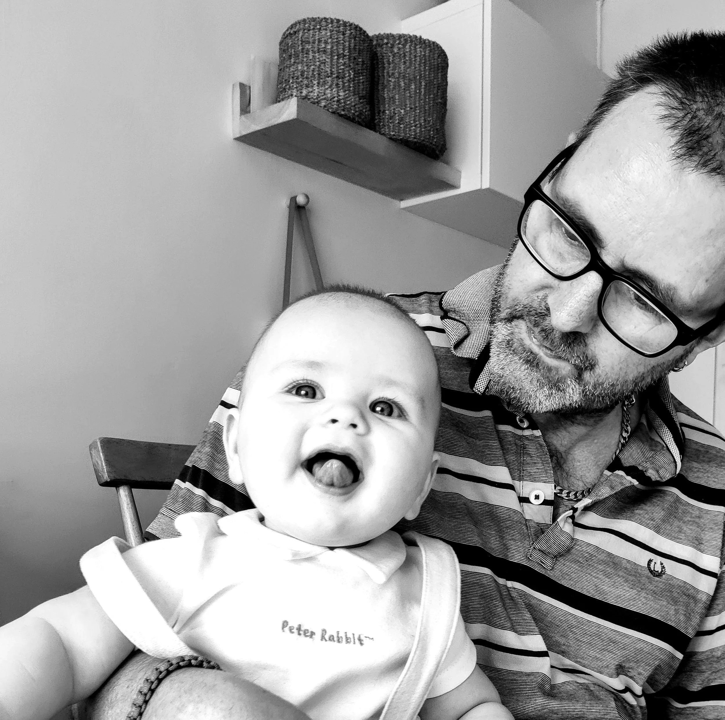 A man with glasses and a beard holding a baby with a big smile and tongue out in a home setting with shelves and baskets in the background.