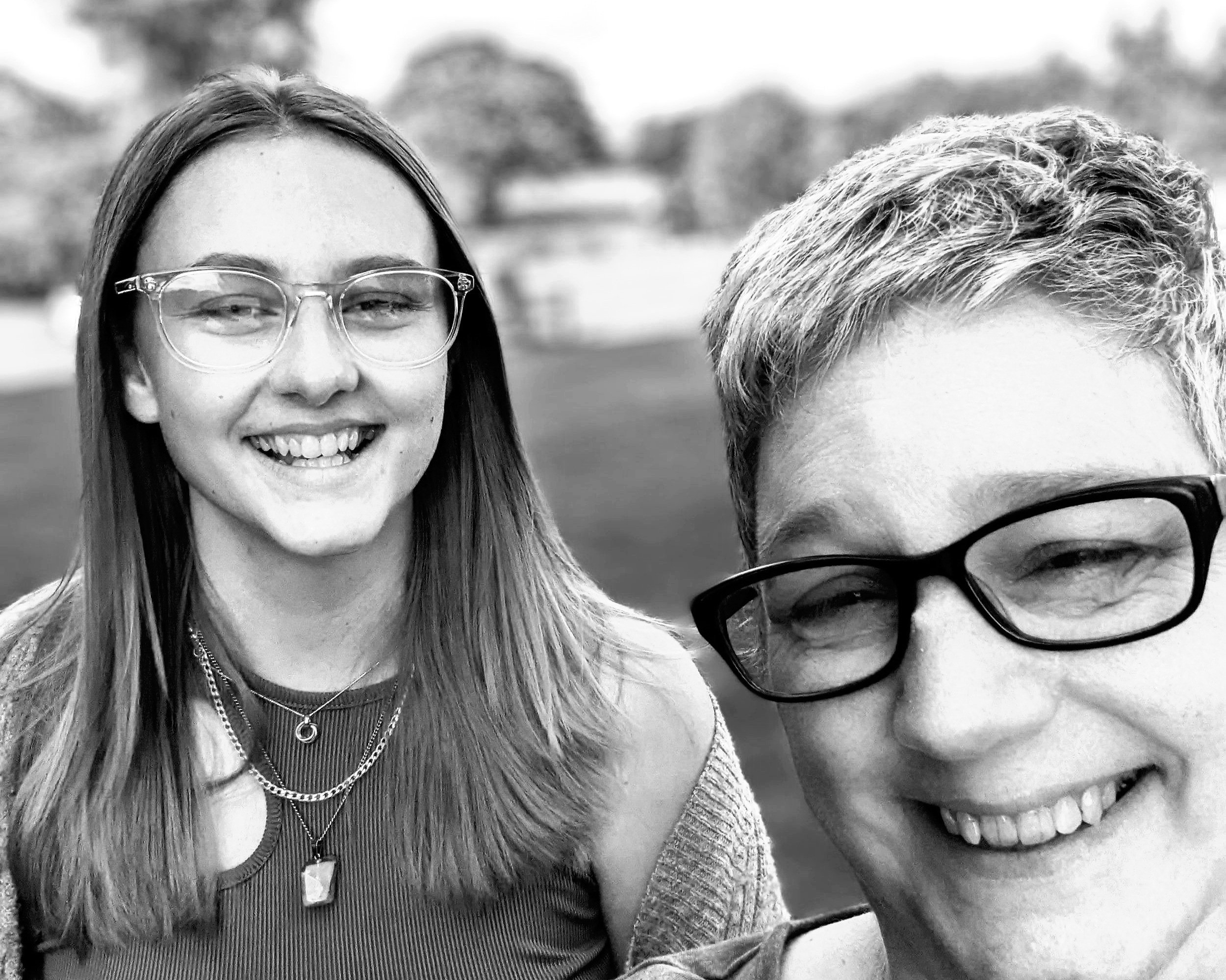 Black and white photo of a smiling woman with long hair, glasses, and layered necklaces, and a smiling person with short hair and glasses in the foreground, outside with trees and a field in the background.