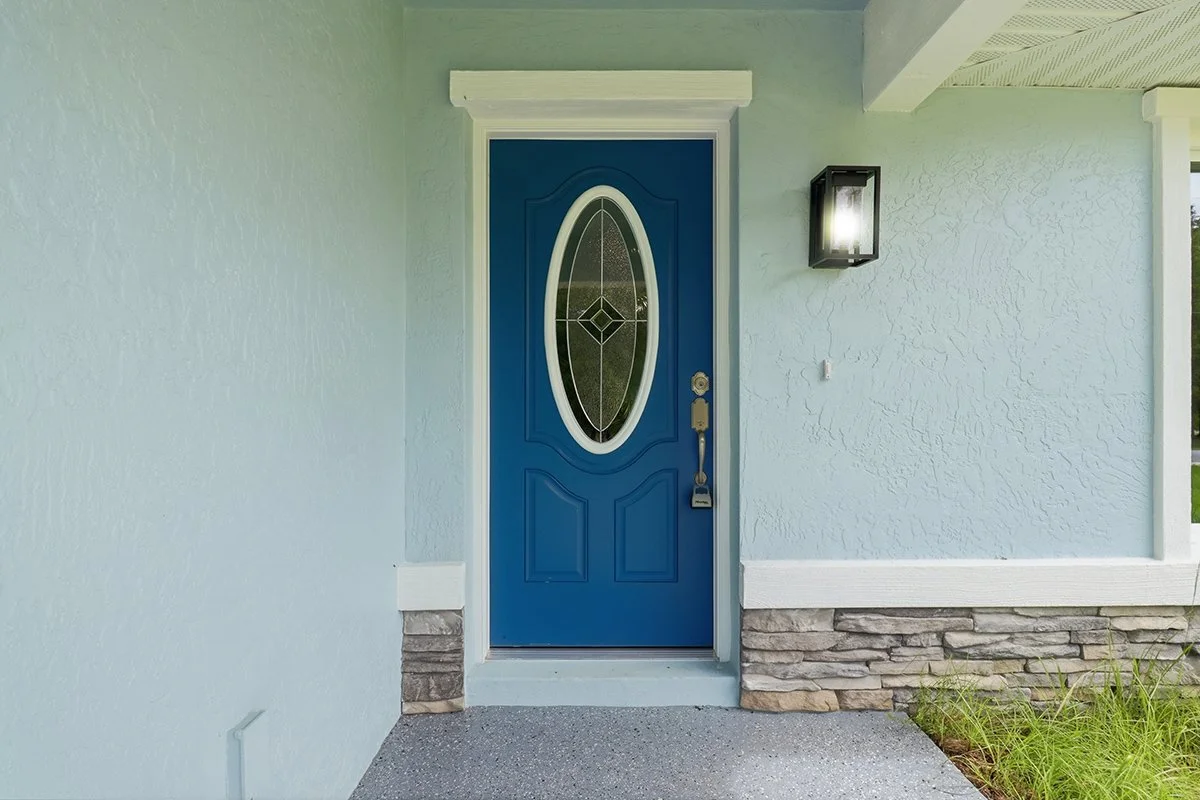 Front door with blue exterior and glass oval window, stone foundation, black wall light, and white trim.