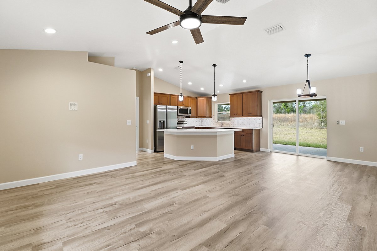 Interior of a modern open-concept living space with a kitchen and dining area, featuring beige walls, wooden cabinets, a ceiling fan, pendant lights, and sliding glass doors leading to an outdoor space.