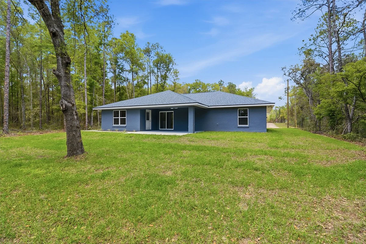 A single-story blue house with a covered patio area, surrounded by a grassy lawn and tall trees under a partly cloudy sky.