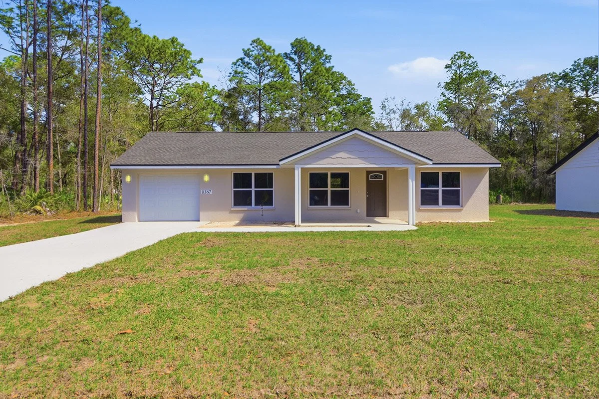 Single story beige homes in Crystal River, Florida with beige shingles, a dark green front door, and a single car garage with a white door. The home has a small, covered front porch, a concrete driveway and sits on a grassy lot.