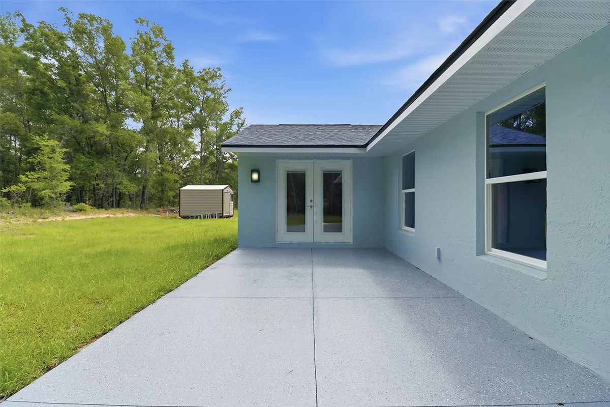 Backyard patio with light gray concrete, white house exterior with two large windows and glass double doors, outdoors green lawn, trees, shed in the background, clear blue sky.