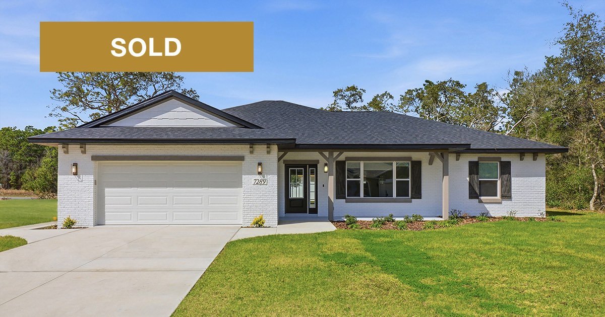 Single-story Citrus Springs home with white painted brick exterior, a charcoal gray shingled roof, large front windows, a two car garage with white door. The homes has gray accents on the corbels and porch beams and a black front door