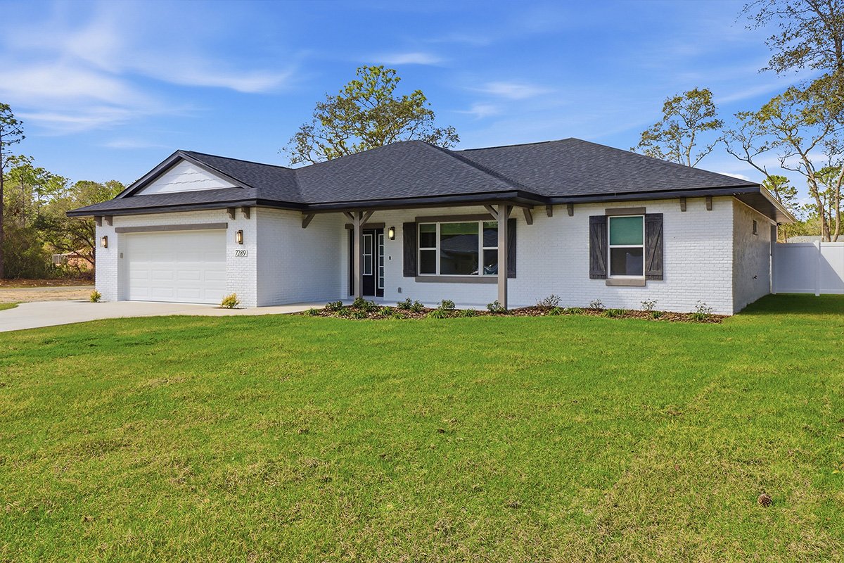Front view of a modern single-story white brick house with black shutters, a black roof, and a two-car garage, surrounded by a green lawn and trees under a blue sky.