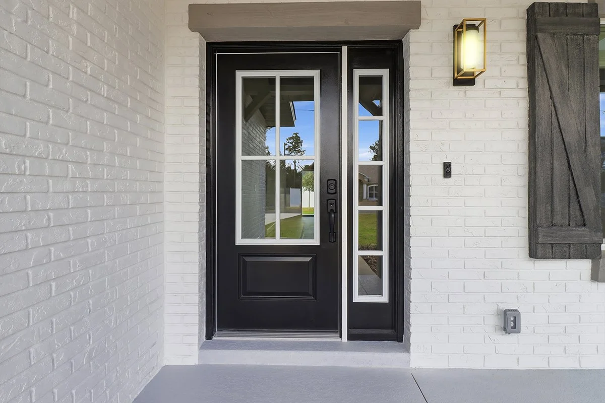 Front door of a house with black frame, divided glass panels, and a black handle, set in a white brick wall with a rustic wooden shutter and a modern wall sconce.