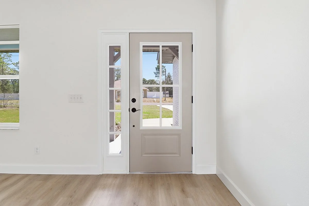 Interior view of a house door with glass panels leading to the outside, with white walls and light wood flooring.