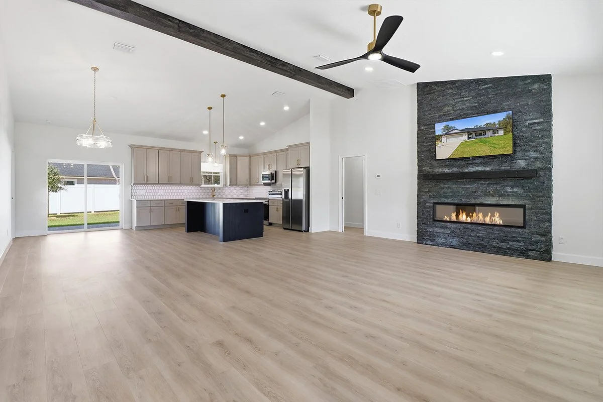 Open-concept living room and kitchen with light wood floors, modern appliances, a stone fireplace with a mounted TV, and large sliding glass doors leading outside.