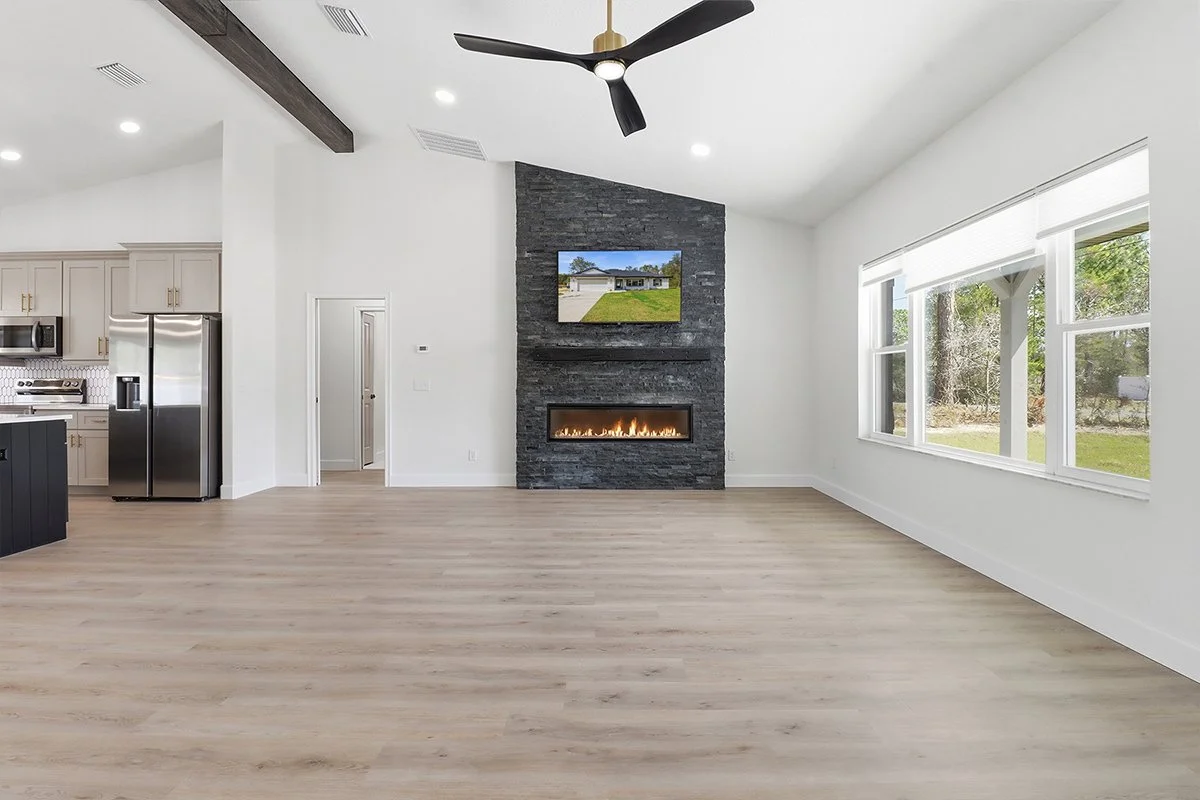Bright living room with large windows, a black ceiling fan, a modern fireplace with a stacked stone surround, and a wall-mounted TV above the fireplace.