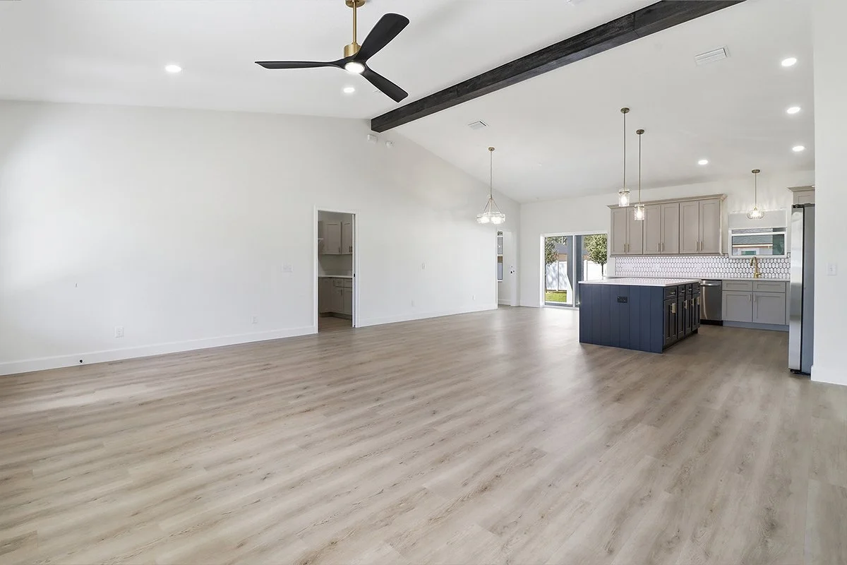 Empty open-concept living room and kitchen with light hardwood floors, white walls, a black ceiling fan, modern light fixtures, a kitchen island painted dark blue, light gray cabinetry, and stainless steel appliances.