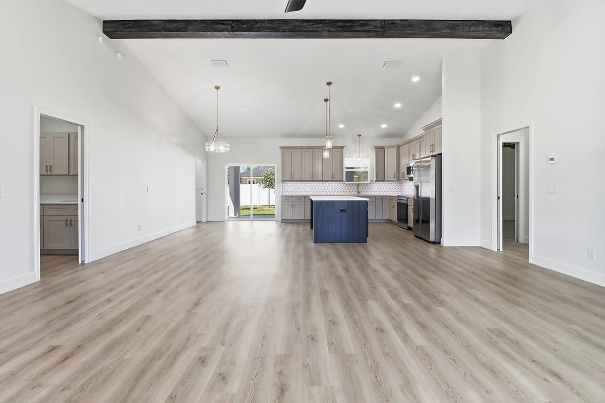 Empty kitchen and living area with light wood flooring, white walls, and gray kitchen cabinets. A kitchen island is painted dark blue, and there are stainless steel appliances. Large windows and sliding glass door provide natural light.