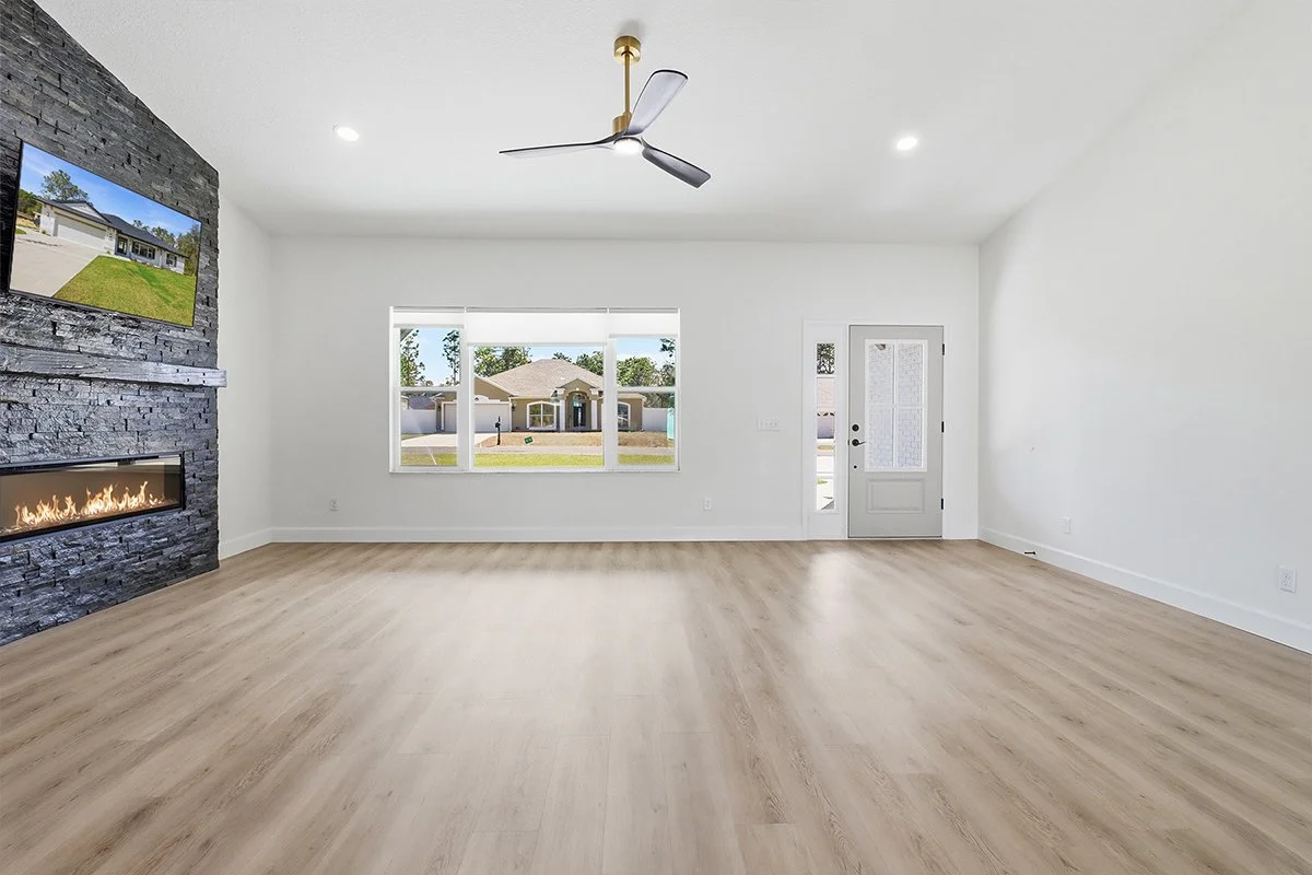 Empty living room with light wood flooring, white walls, a modern ceiling fan, a window looking out to a residential neighborhood, a door with a glass panel, and a stone fireplace with a mounted flat-screen TV.