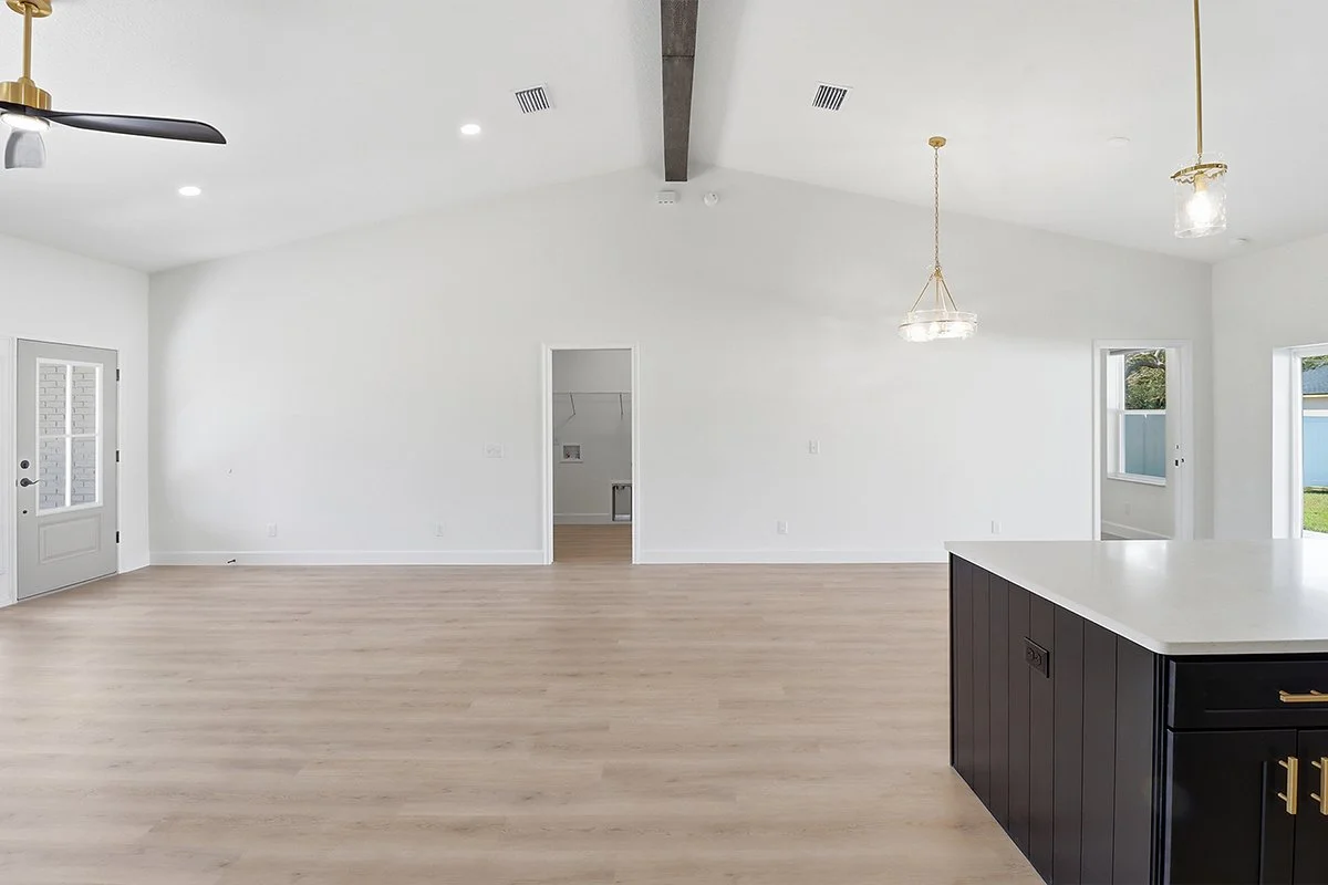 Empty modern living space with white walls, light wood flooring, and large windows. A kitchen island with a white countertop and black base is visible. There are ceiling lights and a ceiling fan.