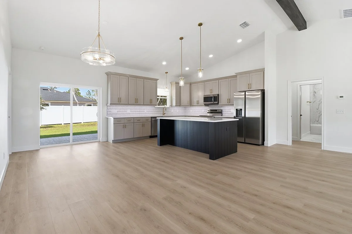 Empty kitchen with light wood flooring, gray cabinets, a black kitchen island, stainless steel appliances, and a sliding glass door leading to a backyard.