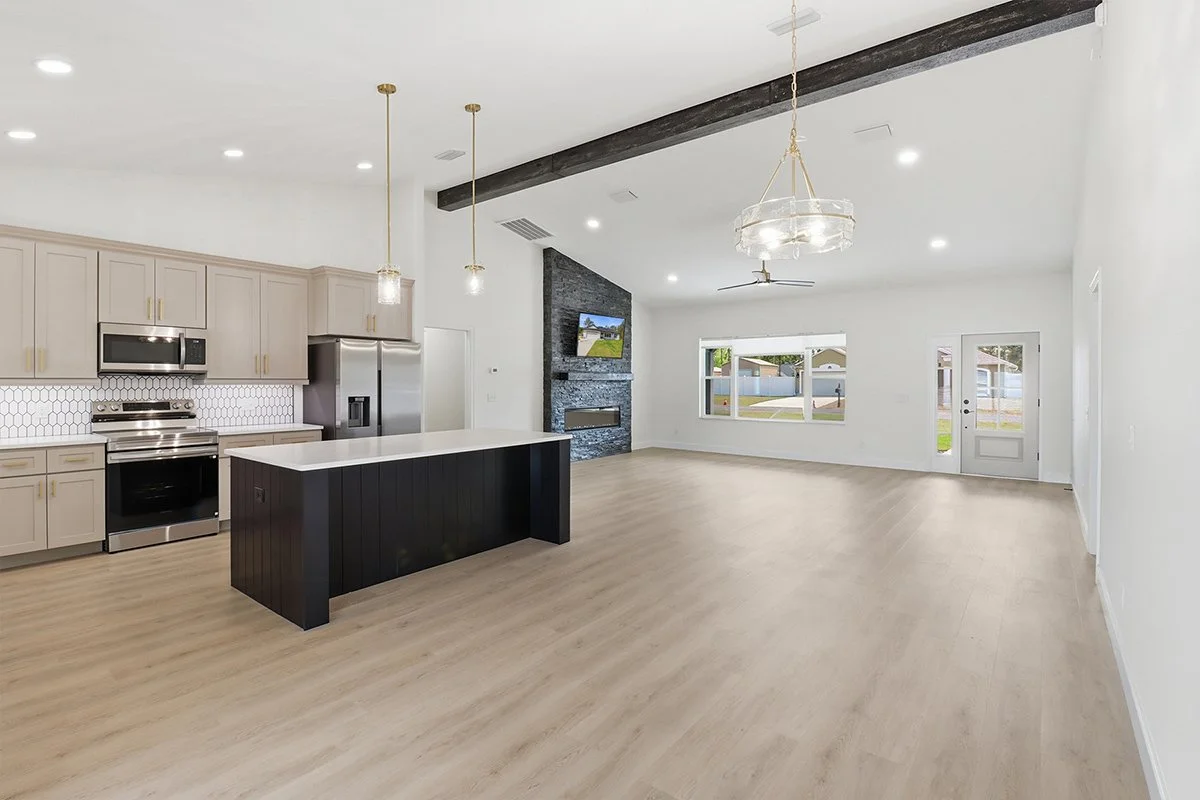 Clean, modern open-concept living room and kitchen with light wood floors, beige cabinets, stainless steel appliances, black island, fireplace, wall-mounted TV, large windows, and ceiling fans.