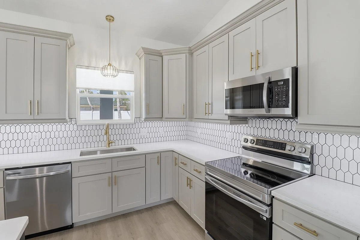 Kitchen with white cabinets, white countertops, honey gold handles, black and white hexagonal backsplash, stainless steel dishwasher, microwave, and oven, window with blinds, and hardwood flooring.