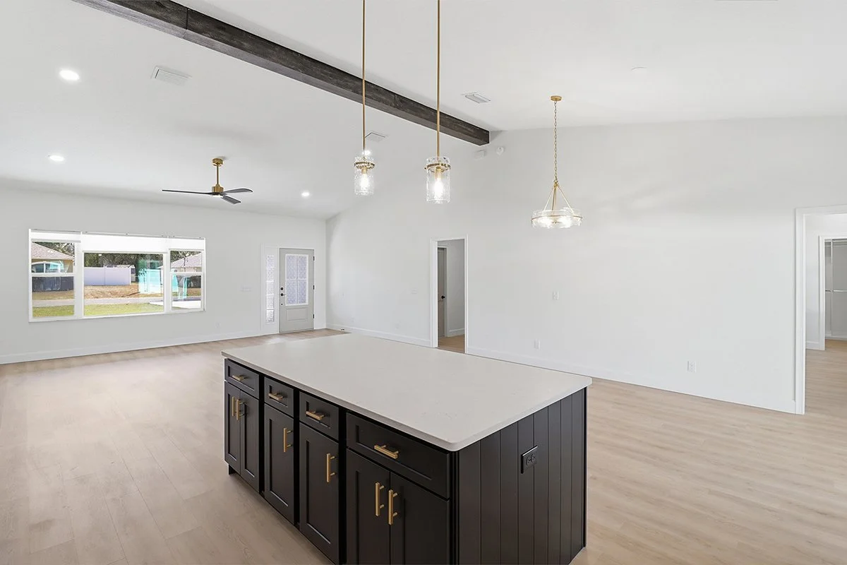 Empty open-concept living and kitchen area with a large island in the foreground, three pendant lights hanging above, large window, white walls, light wood flooring, ceiling fan, and a door leading outside.