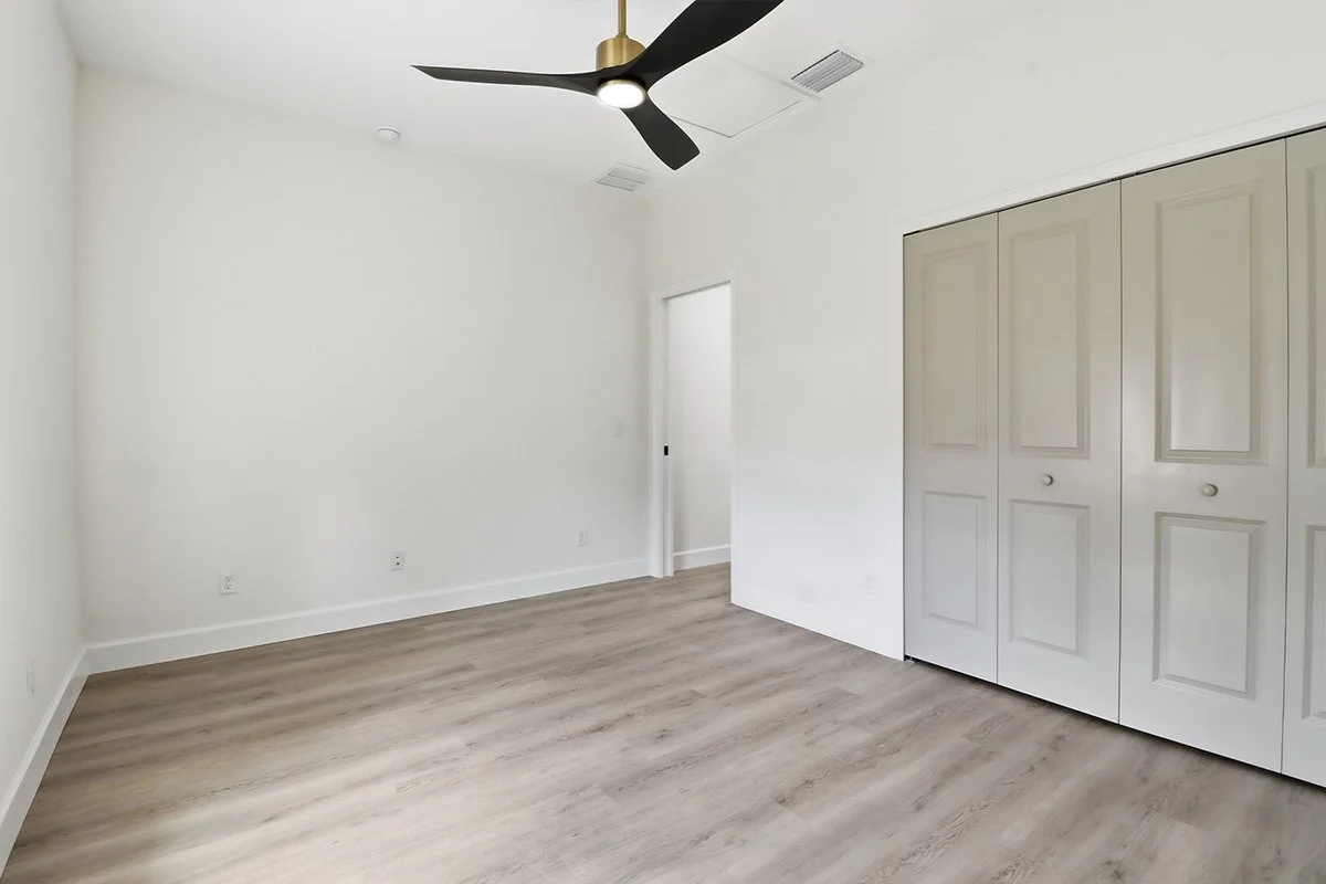 Empty bedroom with white walls, light wood flooring, a closet with double doors, a ceiling fan, and a door leading to another room.