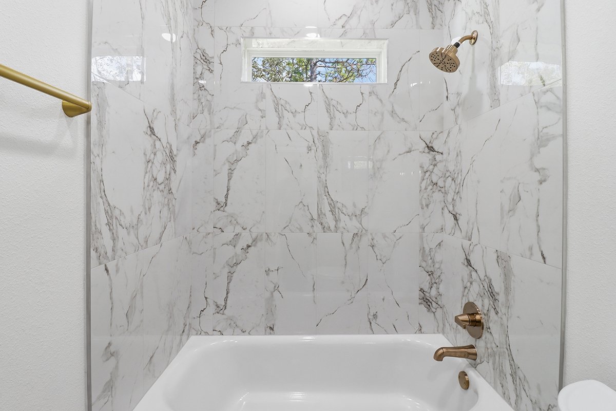 A modern bathroom shower with marble tile walls, a small rectangular window, a gold showerhead, and gold control fixtures.