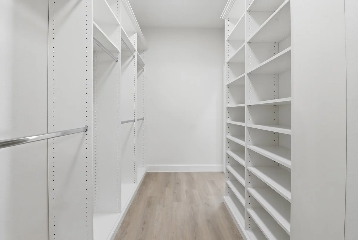 Empty walk-in closet with white shelving, rods for hanging clothes, and light wood flooring.