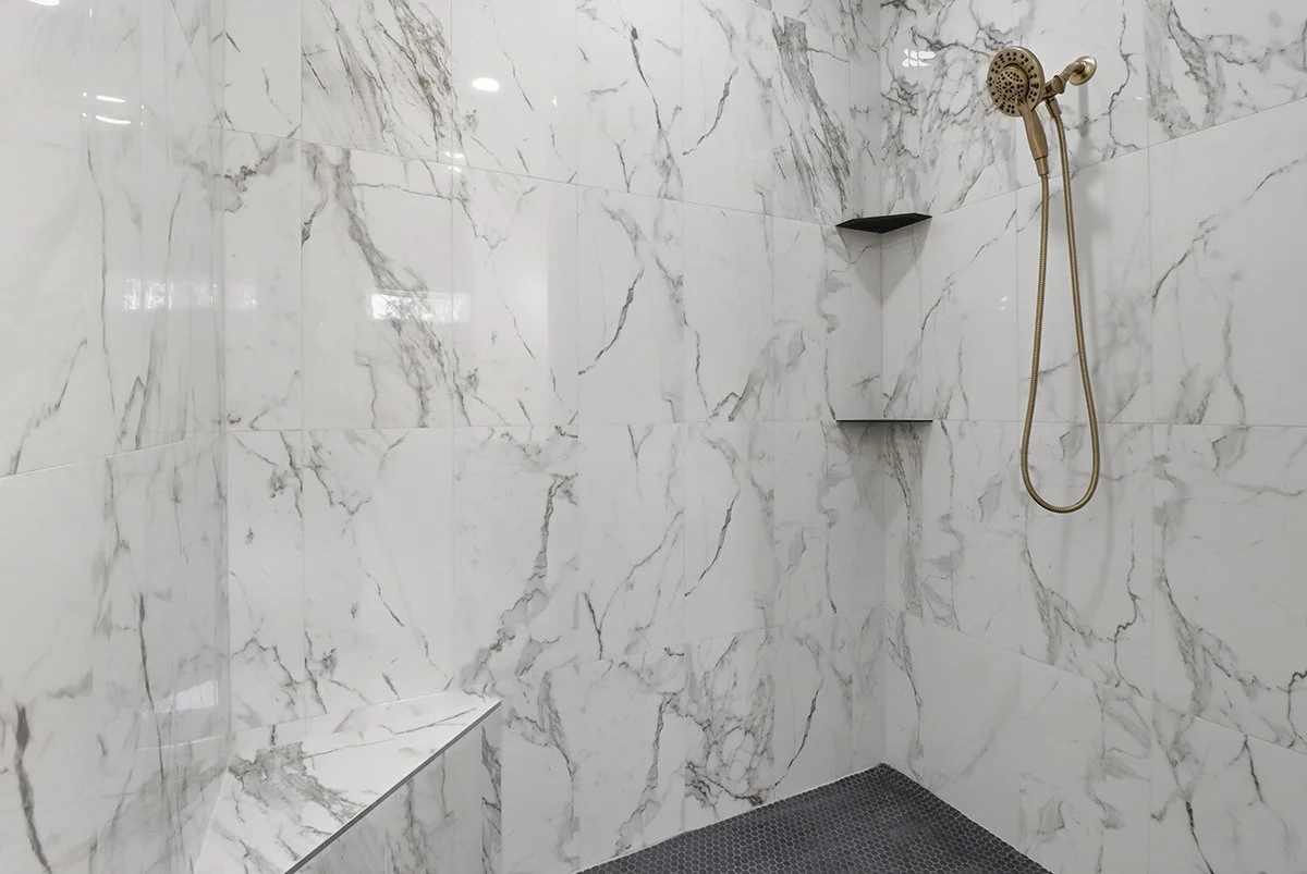 A modern shower with white marble tiles with gray veining, a built-in corner bench, a black corner shelf, and a brass shower head with a hose.