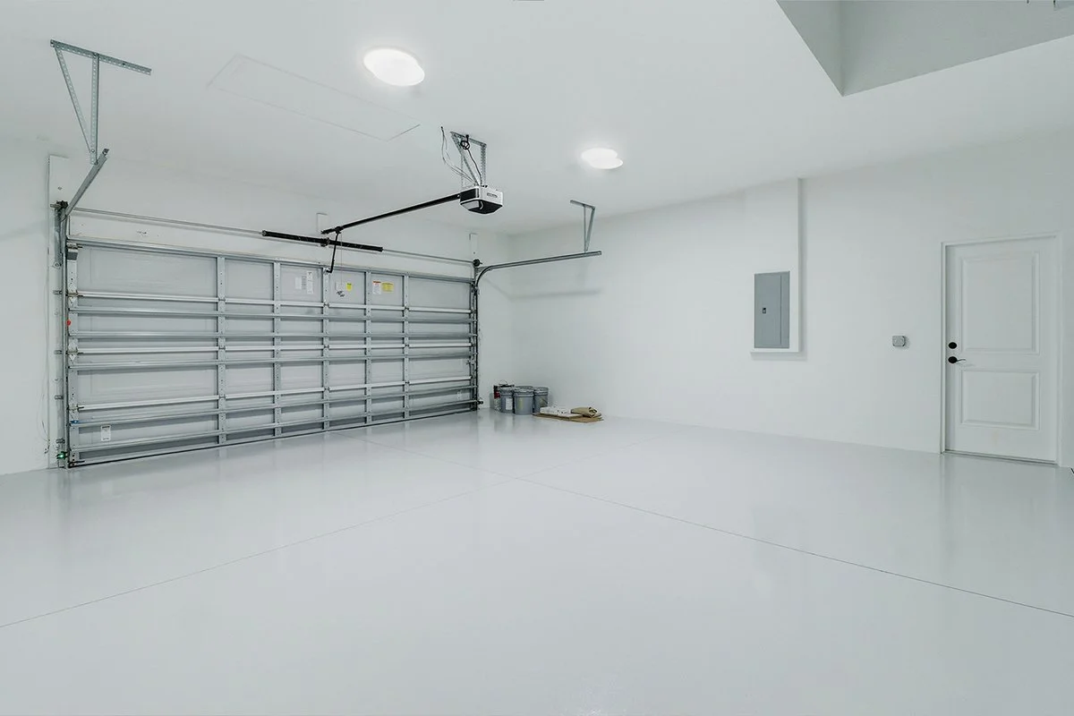 Empty white garage with a closed metal sectional door, overhead lighting, a small electrical panel on the wall, and a door on the right side.