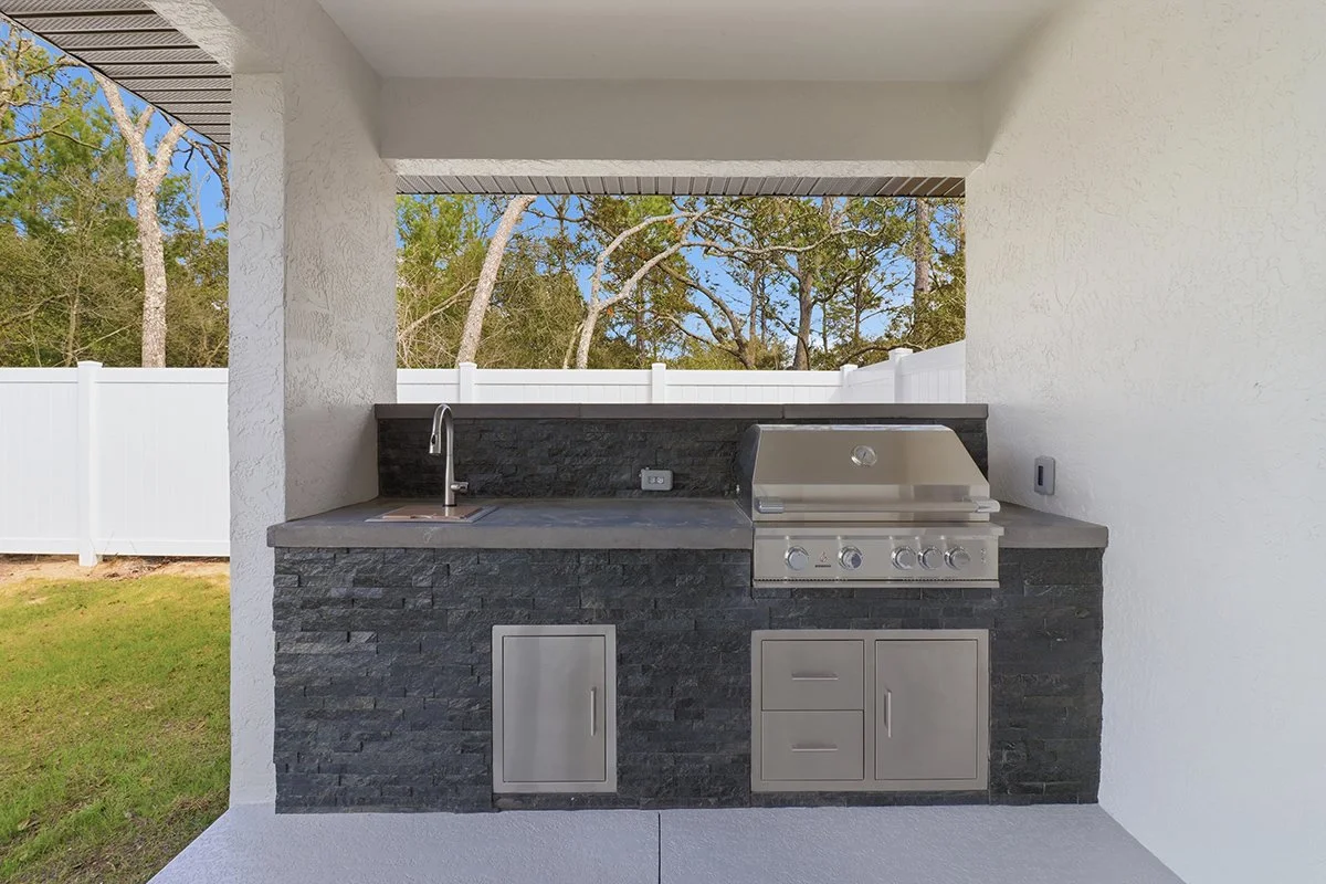 Outdoor kitchen area with a built-in stainless steel grill, a small sink, black stone counter and cabinets, and a white fence in the background.