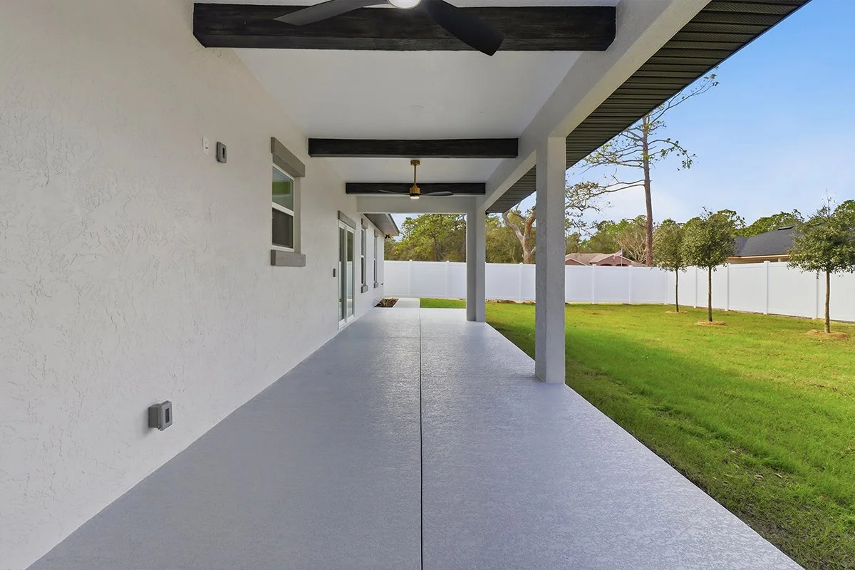 Empty covered patio with white stucco wall, small windows, ceiling fans, and a view of a backyard with green grass, small trees, and a white fence