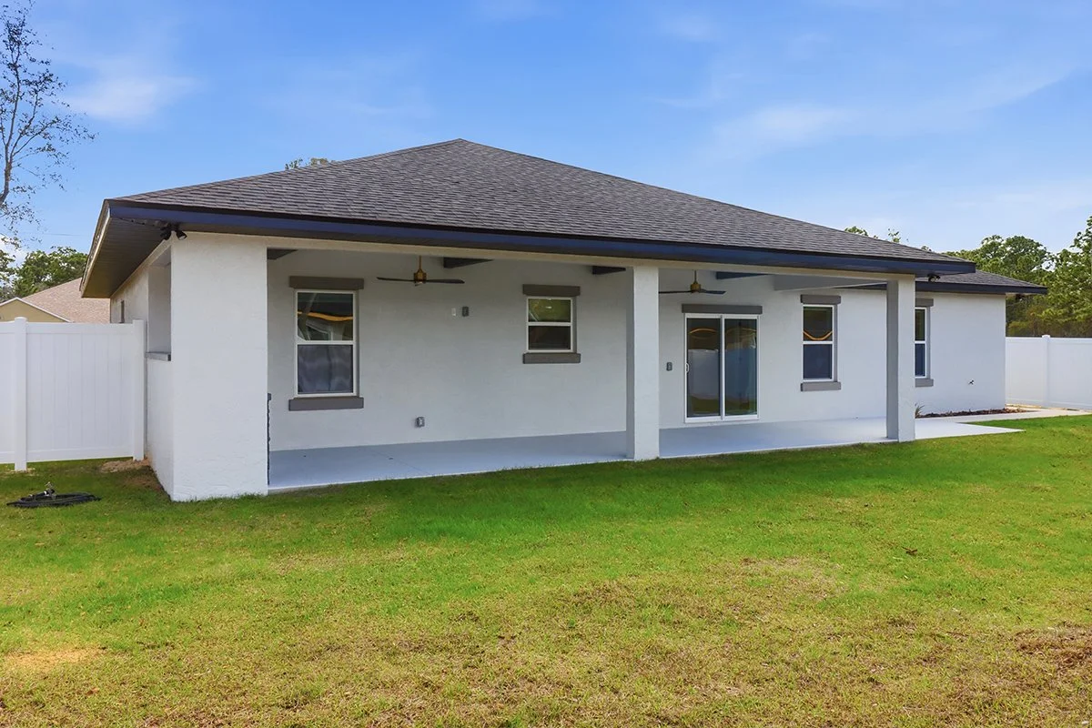 Rear view of a modern white house with a dark gray roof, three small windows, and a sliding glass door, surrounded by a green lawn and white fence.