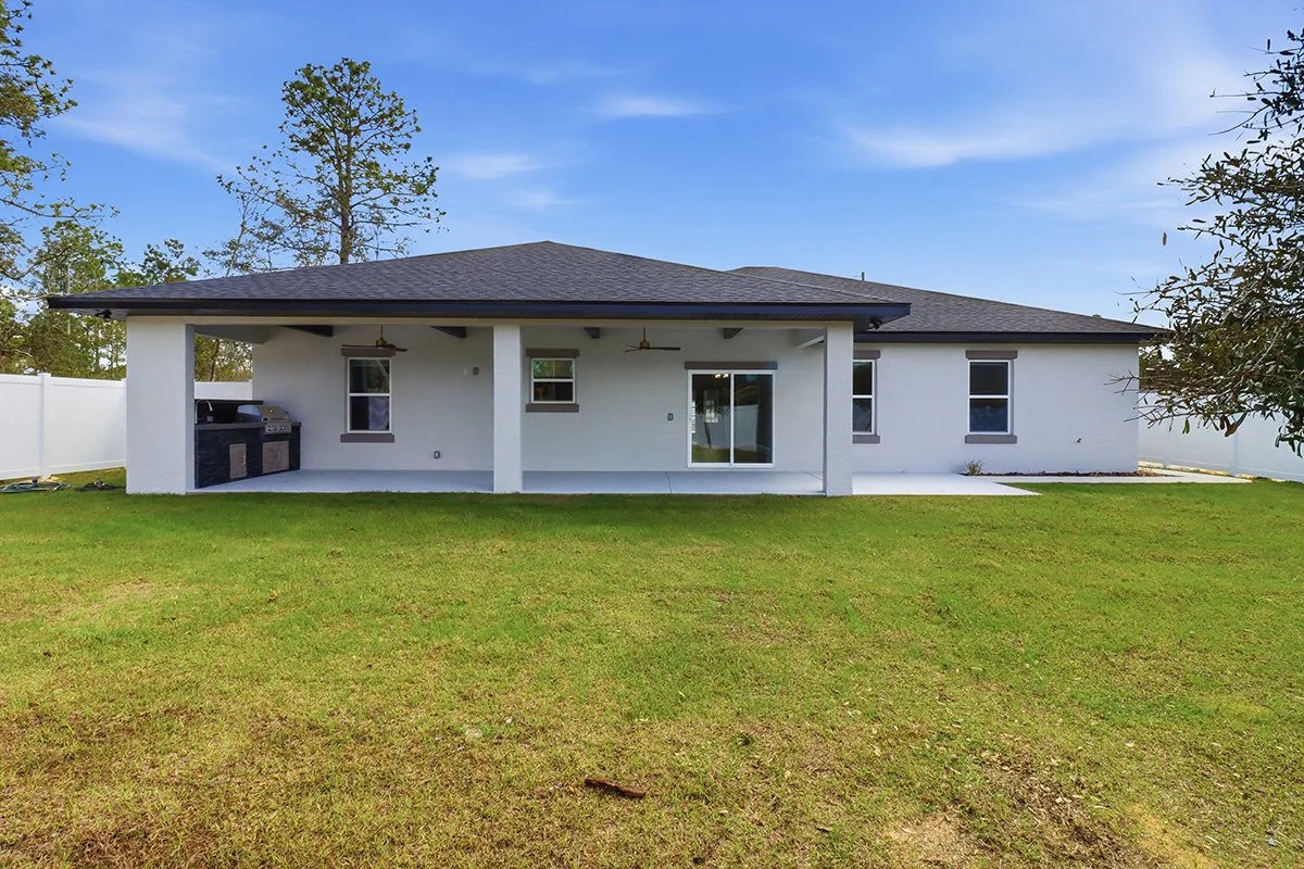 Rear view of a modern, single-story house with a covered patio, a grill, and a fenced backyard with green grass.