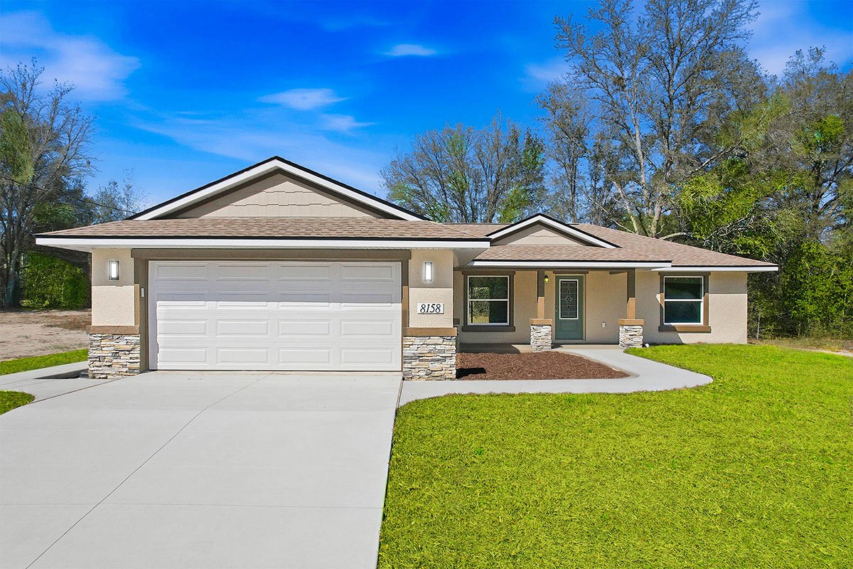 Newly constructed beige home with green front door and beige shingles. The house has a white 2-car garage, with concrete driveway and sidewalk leading to front door. Home sits on a grassy lot with mature trees in backyard.