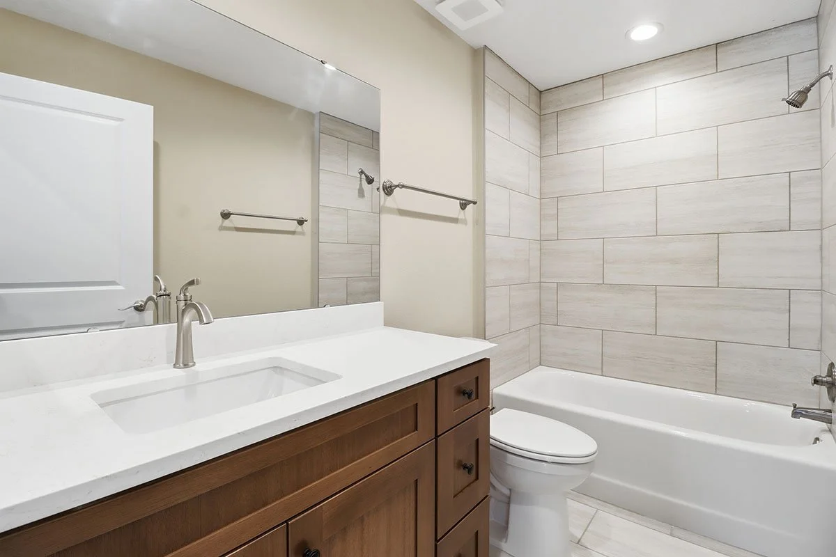 Modern bathroom with a wooden vanity, white countertop, a built-in sink, a large mirror, a toilet, a bathtub, and tiled walls.