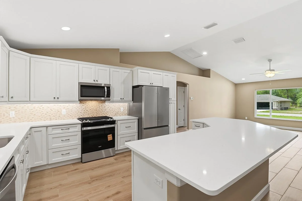 Modern kitchen with white cabinets, a large island with a white countertop, stainless steel appliances including a refrigerator, microwave, and oven, and a window overlooking a suburban neighborhood.