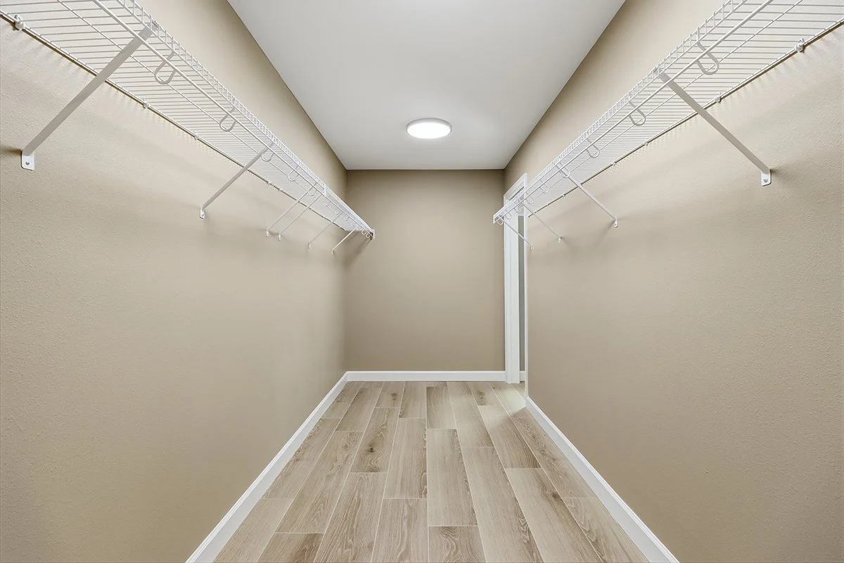 Empty walk-in closet with beige walls, white wire shelves on both sides, and light-colored wooden flooring.