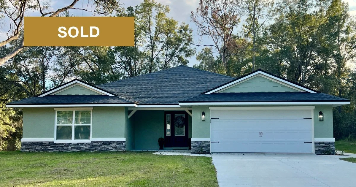 Home with pale green stucco exterior, black and grey stone accents, black shingles, two car garage, covered front porch and front window opposite the garage