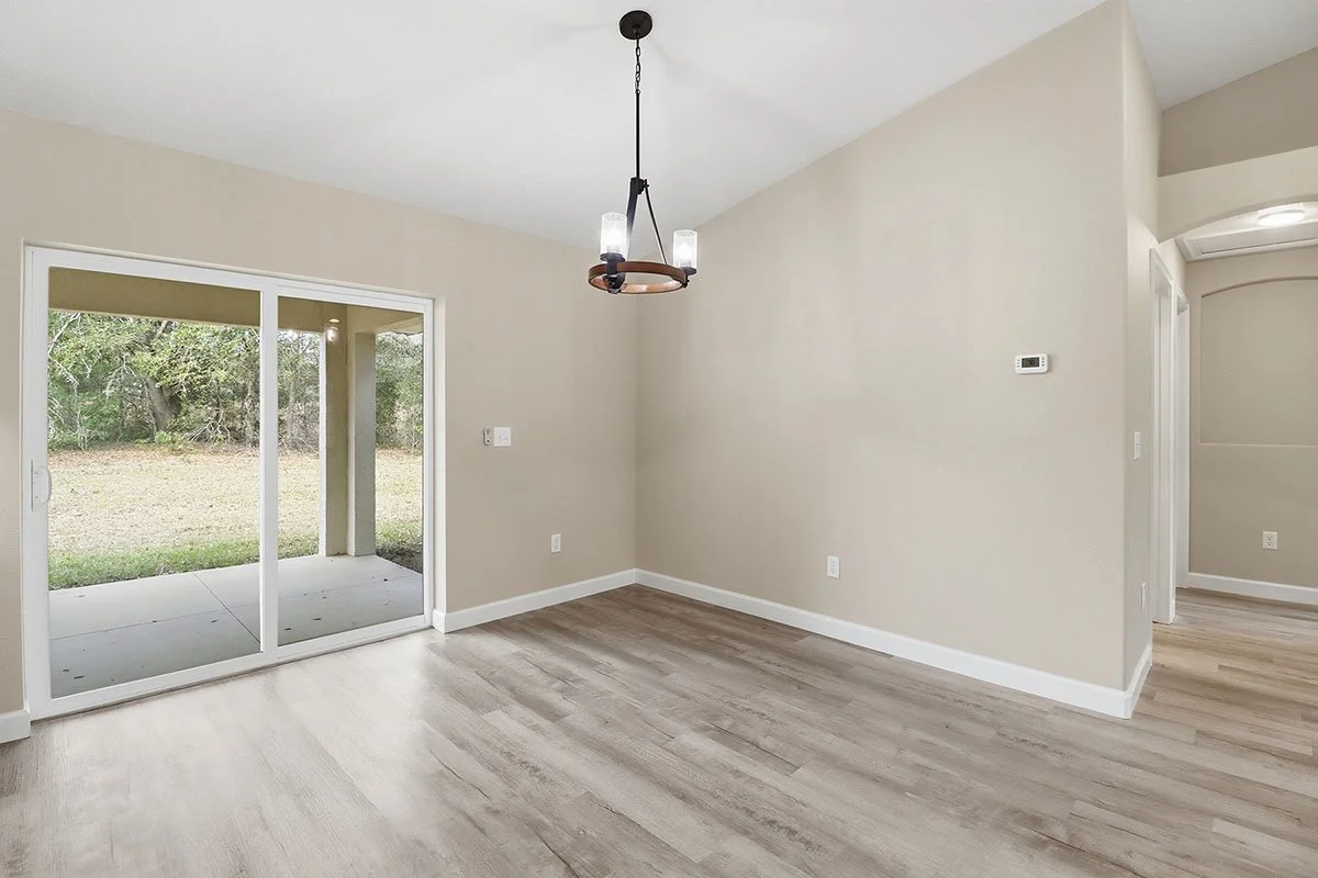 Empty living room with sliding glass door leading to backyard, beige walls, wood-look flooring, and a modern chandelier hanging from the ceiling.