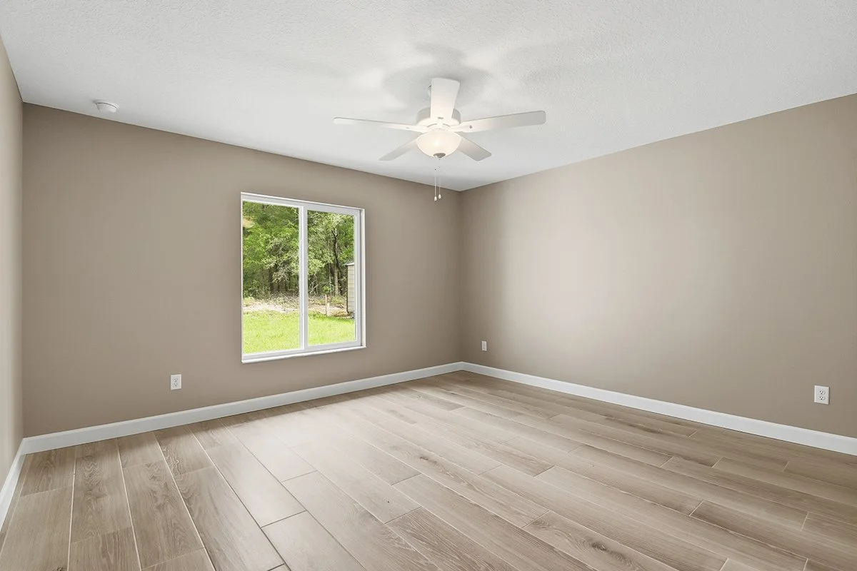 Empty room with beige walls, light wood flooring, a window showing trees outside, and a ceiling fan with a light.