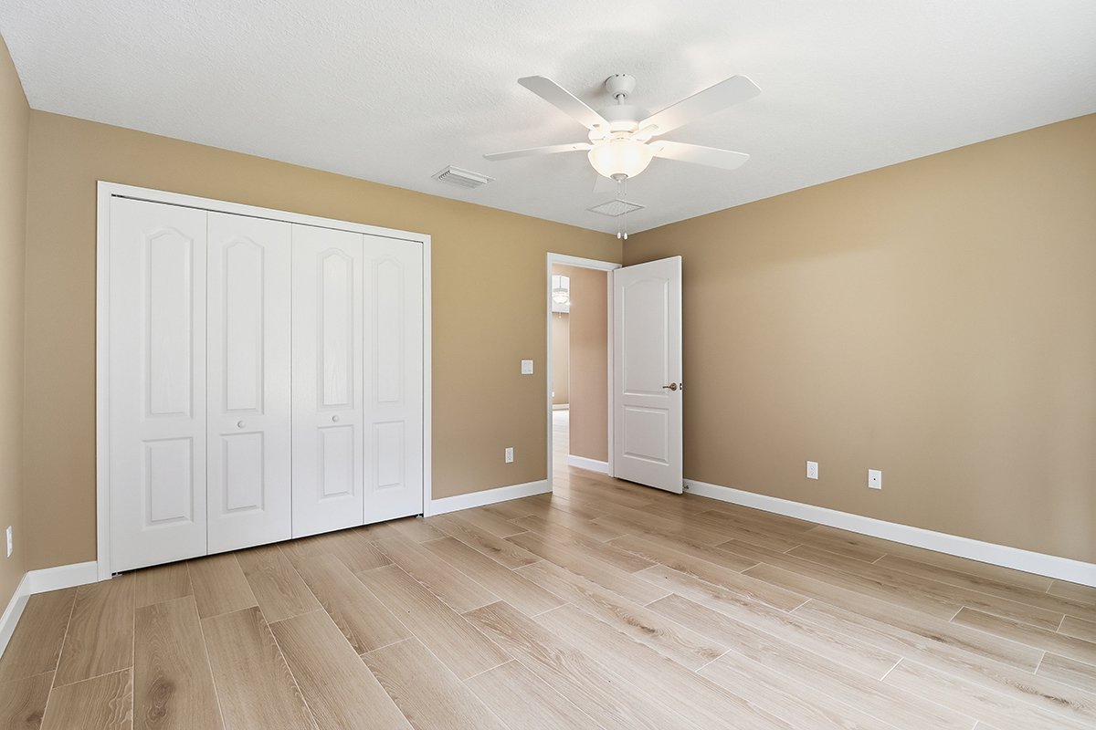Empty bedroom with beige walls, white closet doors, hardwood floor, open door leading to another room, ceiling fan, and ceiling vent.