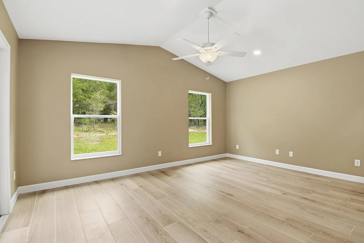 Empty room with beige walls, light wood flooring, two windows showing greenery outdoor, white ceiling fan, and multiple electrical outlets.