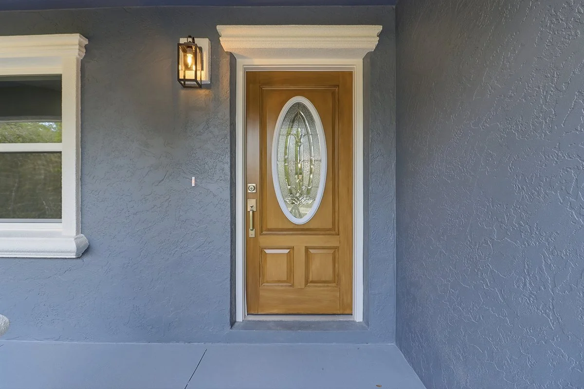 Front porch of a house with a wooden door featuring an oval glass window, a black lantern-style light fixture, a gray textured wall, white trim, and a window to the left.