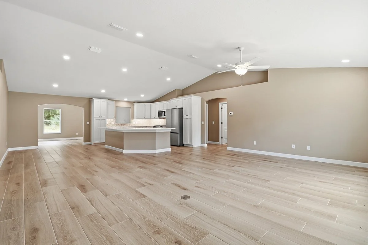 Empty open-concept living room and kitchen with beige walls, white cabinets, stainless steel appliances, and light wood flooring.