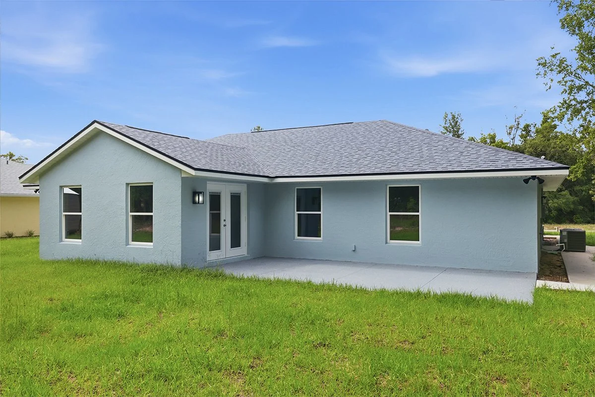 A newly built, single-story house with light blue exterior walls, a gray shingle roof, and a small backyard. The house has three windows, a glass door with side windows, and outdoor wall-mounted lights. The yard features green grass, and there are ne