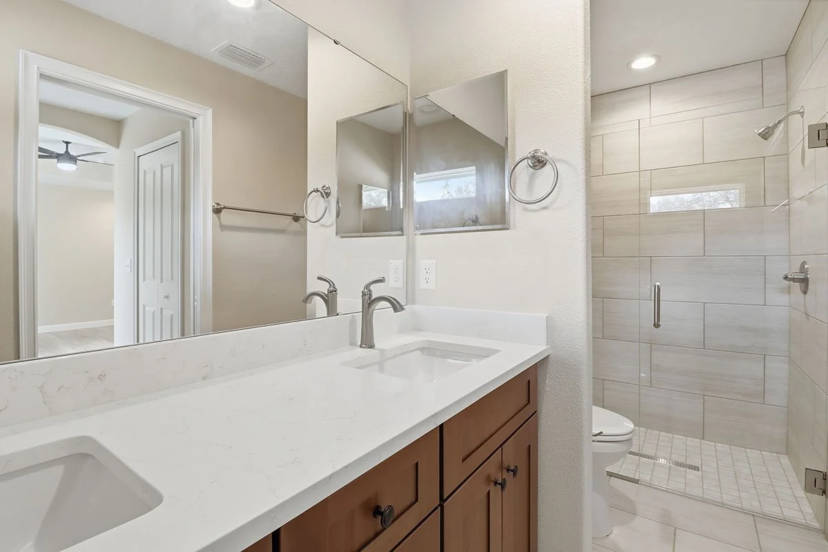 Bathroom with double sink vanity, mirror, towel rings, and a glass-enclosed shower with beige tile walls.