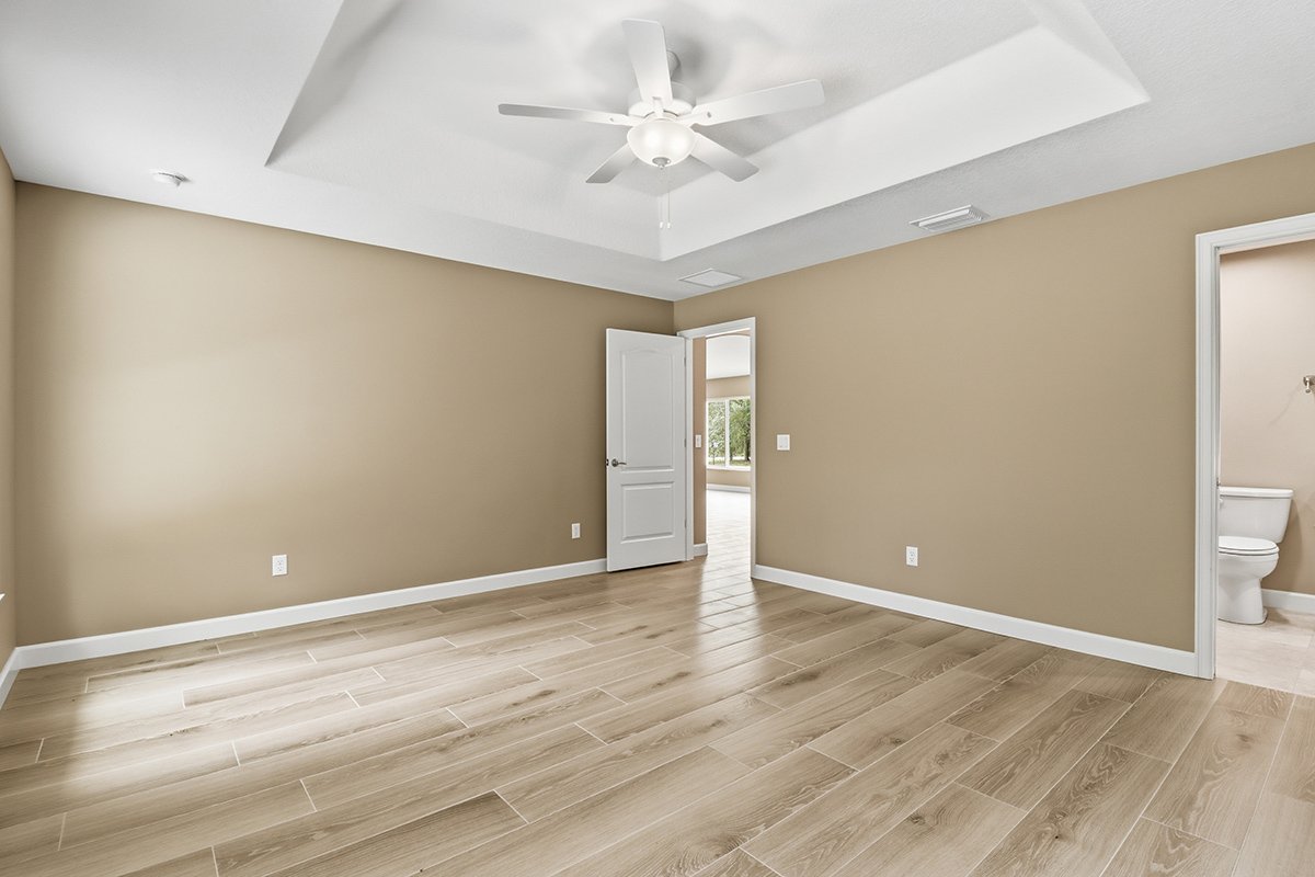 Empty room with beige walls, wood flooring, ceiling fan, open door to another room, and a bathroom with a toilet.