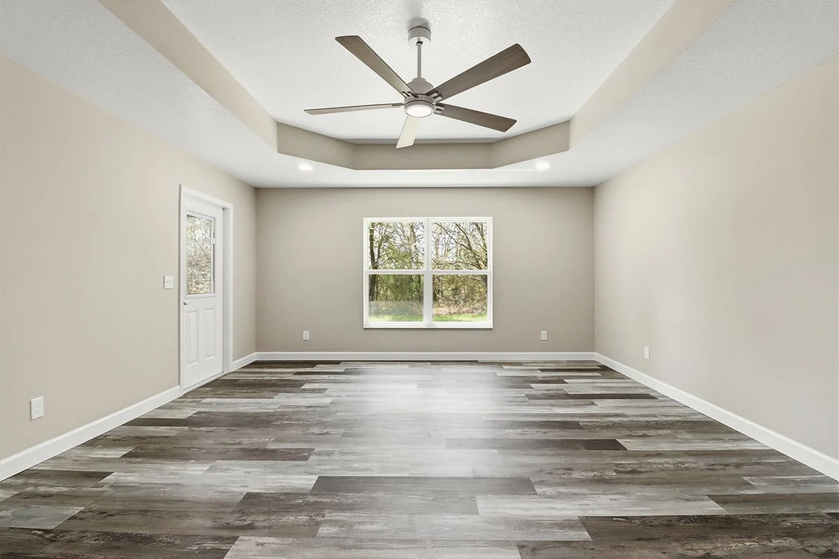 Empty living room with beige walls, wood flooring, a ceiling fan, a window showing a tree outside, and a door leading outside.