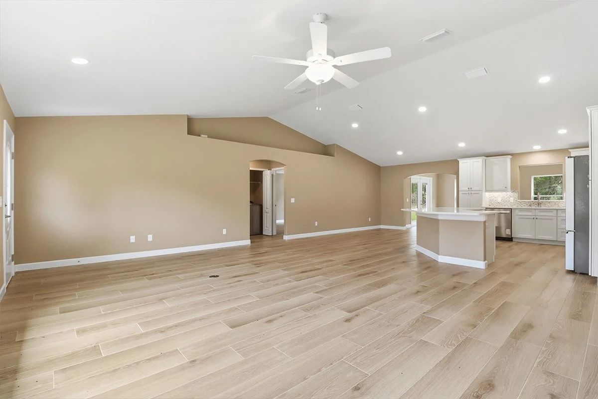 Empty living room and kitchen with wood flooring, beige and white walls, white ceiling, ceiling fan, and natural light from windows.