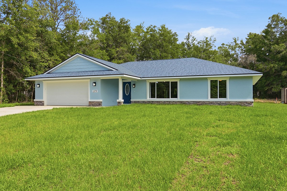 A newly built single-story house with a blue exterior, white garage door, stone accents, and a front lawn, with trees in the background and a partly cloudy sky.