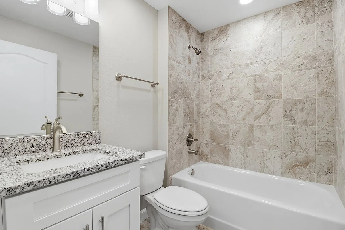 Bathroom with granite countertop sink, white cabinet, toilet, and bathtub with beige tile walls.