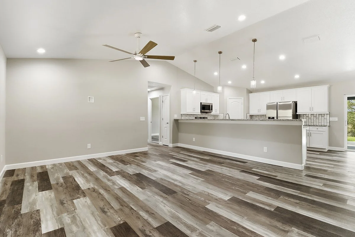 Empty living room with wood plank flooring, ceiling fan, and an open kitchen with white cabinets, stainless steel appliances, and pendant lighting.