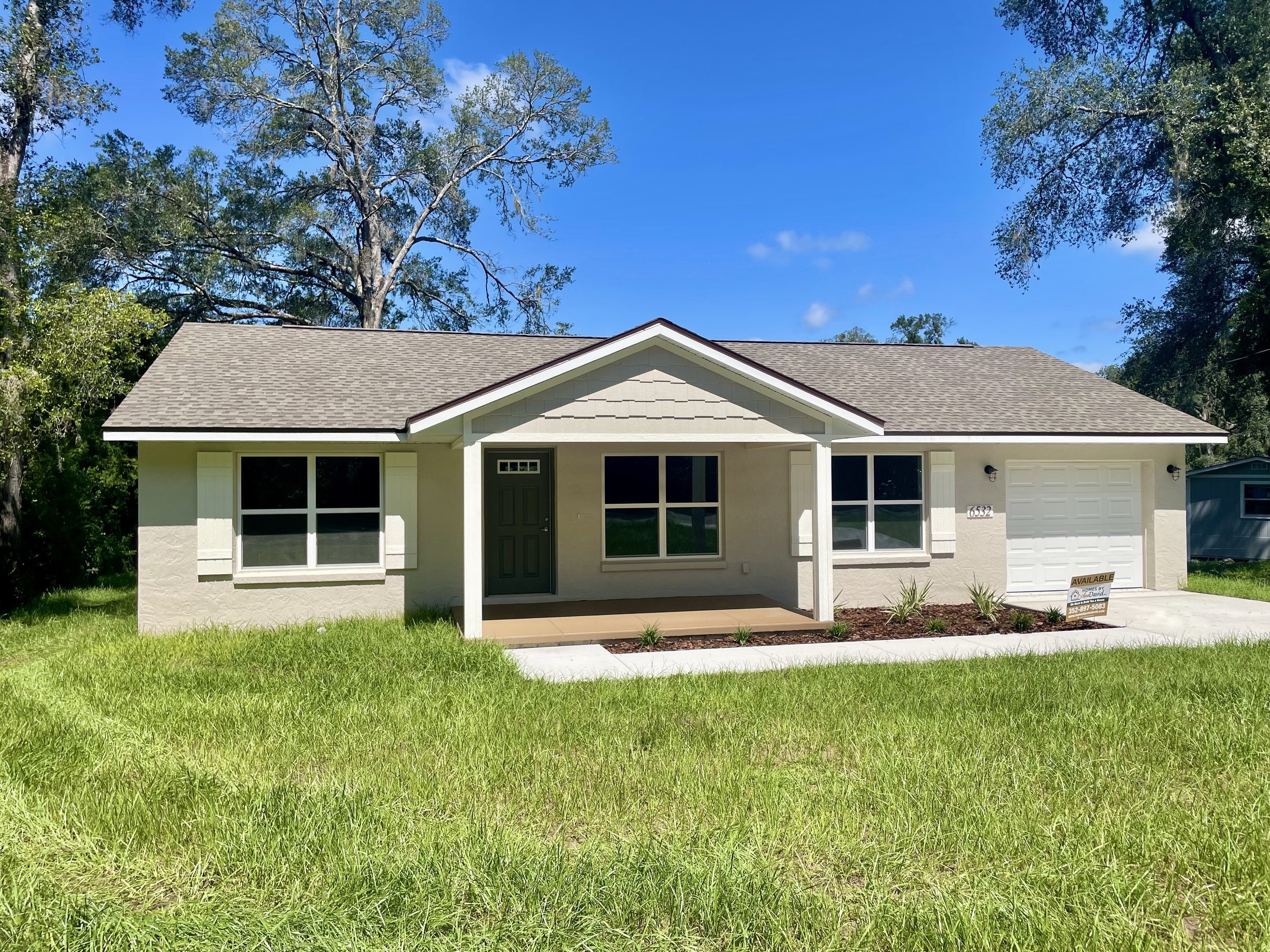 Newly constructed home with a beige exterior and dark green front door with small front porch, white shutters on windows, and a single car garage with white door. It has a concrete driveway with sidewalk to the front porch and green grass.